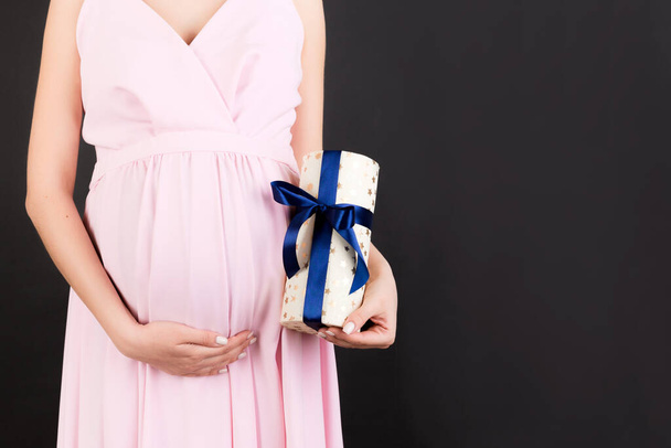 Cropped image of pregnant woman in pink dress holding a gift box and touching her belly at black background. Expecting a baby boy. Copy space. - Photo, Image