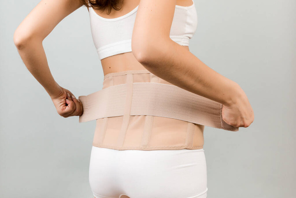 Back view of pregnant woman in underwear putting on supporting bandage to reduce backache at gray background with copy space. Cropped image of orthopedic abdominal support belt concept. - Photo, Image
