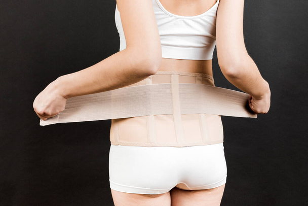 Back view of pregnant woman in underwear dressing orthopedic corset to make the backache go away at black background with copy space. Close up of orthopedic abdominal support belt concept. - Foto, imagen