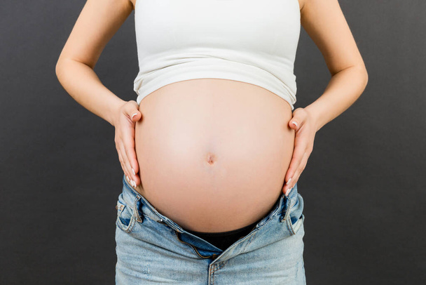 Close-up van zwangere buik op kleurrijke achtergrond met kopieerruimte. Toekomstige moeder in geopende jeans omarmt haar naakte babybuil. Moederschap concept. - Foto, afbeelding