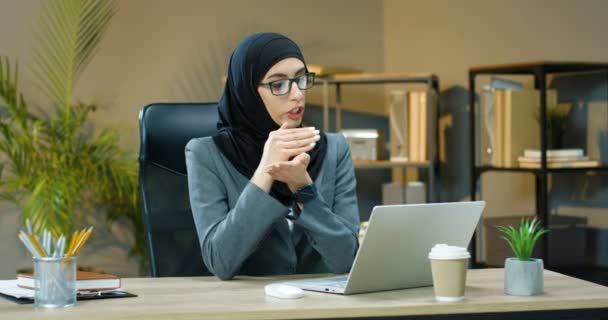 Muslimi nuori nainen hijabissa ja lasit istuu toimistossa ja puhuu webbikameran kautta kannettavalla tietokoneella. Kaunis arabialainen liikenainen perinteisessä huivissa, jolla on videochat tietokoneella. Videolähetys. - Materiaali, video