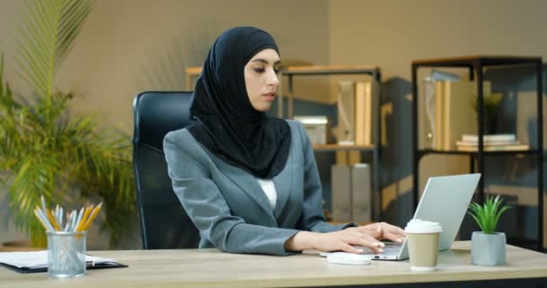Portrait shot of young Arabic woman in hijab scarf sitting at table in office, working on laptop and smiling to camera. Female muslim businesswoman typing on keyboard of computer at desk and smile. - Footage, Video