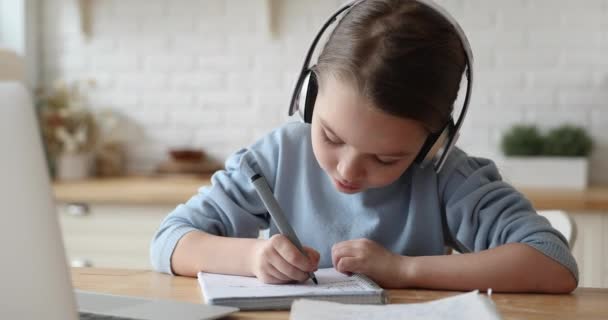 Klein basisschoolmeisje met koptelefoon, huiswerk maken op afstand. - Video