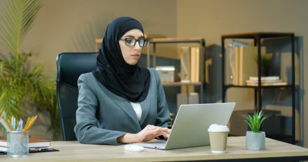 Portrait of young Arabic woman in hijab and glasses sitting at table in office, working on laptop and smiling to camera. Female muslim businesswoman typing on keyboard of computer at desk and smile. - Footage, Video