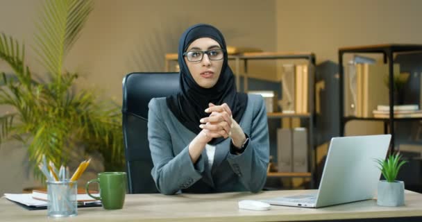 Mladá krásná muslimka v hidžábu a brýle sedí u stolu v kanceláři a mluví s kamerou, jako by něco říkala. Ženské arabské obchodní vlogerky. Online webinář. U telefonu obchodnice. Vlog - Záběry, video