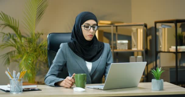 Nuori arabinainen hijabissa ja lasit toimistossa ja läppärissä. Nainen muslimi kirjoittamalla näppäimistöllä tietokoneen ja siemaillen kuumaa juomaa pöydässä kaapissa. Kahvin tai teen juominen. - Materiaali, video