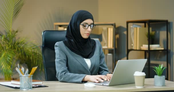 Beautiful young Arabic woman in black hijab and glasses sitting at desk in office and working on laptop. Female muslim manager typing on keyboard of computer at table. Coffee to-go. Arab businesswoman - Materiaali, video