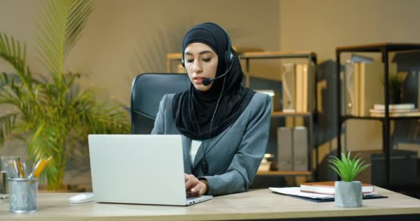 Young muslim woman in black hijab and headset talking via webcam at laptop computer with client in office. Arabian call cener female manager speaking with customer in videochat. Videochatting. - Πλάνα, βίντεο