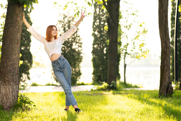 Młoda ruda kobieta w białej bluzce i niebieskich dżinsach stojąca w letnim parku ciesząca się ciepłym dniem. - Zdjęcie, obraz