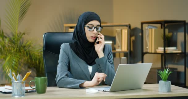 Young Arabic woman in hijab and glasses sitting at desk in office, talking on mobile phone and working on laptop. Female muslim typing on keyboard of computer, speaking on cellphone Arab businesswoman - Filmati, video