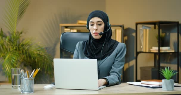 Portrait of young Arabic woman in hijab and headset sitting at table in office, working on laptop and smiling to camera. Female muslim businesswoman typing on computer at desk in call center. - Záběry, video