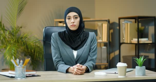 Siyah tesettürlü genç müslüman kadın portresi rahat bir ofiste masada oturuyor ve kameraya mutlu bir şekilde gülümsüyor. Masadaki gülümseyen, oldukça neşeli Arap iş kadını.. - Video, Çekim
