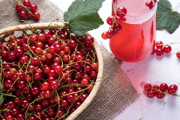 Сприги ягод красной смородины лежат в плетеной тарелке, Морс ягод в стеклянном стакане на светлом фоне - Фото, изображение
