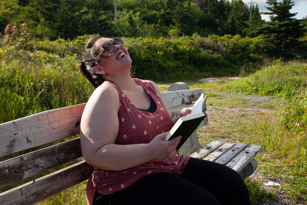 Woman chuckles while reading her book outside on a park bench  - Photo, Image