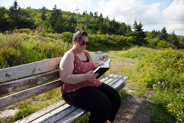 Person lächelt, während sie auf einer Parkbank in der Sonne ein Buch liest - Foto, Bild