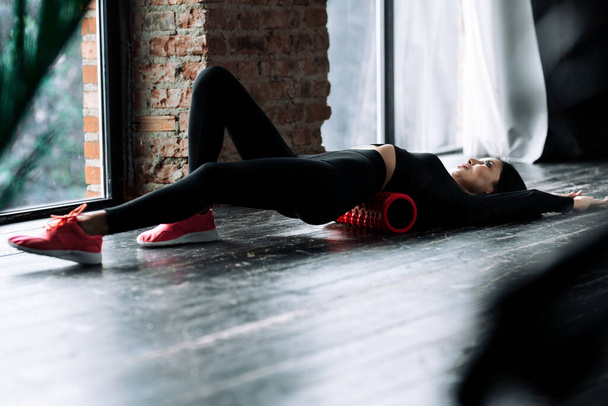 A slender girl in a black sports uniform stretched her legs forward and massages the fascia of the lumbar muscles with a foam roller - Foto, Imagen