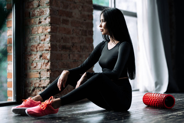 Portrait of a girl in a black sports uniform and with long hair sitting on the floor in the gym who massaged the fascia of the body muscles with a foam roller - Fotoğraf, Görsel
