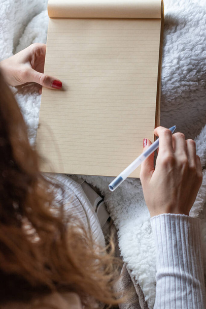 Νεαρή γυναίκα αρχίζει να γράφει σε ένα κενό σημειωματάριο κάθεται στον καναπέ στο σπίτι με ένα στυλό. Το σημειωματάριο είναι κενό. - Φωτογραφία, εικόνα
