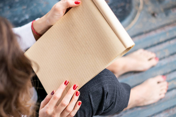 Νεαρή γυναίκα διαβάζει ένα σημειωματάριο καθισμένη ξυπόλητη στο πάτωμα. Έχει βαμμένα νύχια και καστανά μαλλιά. Το σημειωματάριο είναι κενό. - Φωτογραφία, εικόνα