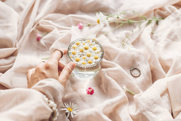 Hand with jewelry holding glass cup with daisy flowers in water on background of soft beige fabric with wildflowers. Tender floral aesthetic. Creative summer image. Bohemian mood - Foto, Imagem
