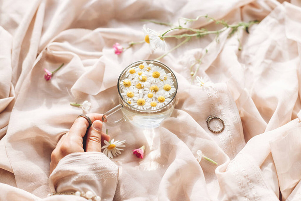 Hand holding glass cup with daisy flowers in water on background of soft beige fabric with wildflowers and jewelry. Tender floral aesthetic. Creative summer image. Bohemian mood. - Foto, Imagem