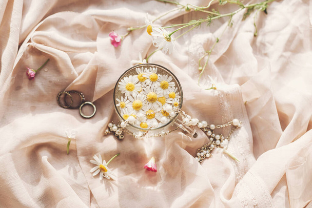 Tender floral aesthetic. Daisy flowers in water in glass cup on background of soft beige fabric with wildflowers and jewelry. Creative summer image, flat lay. Bohemian mood - Photo, image