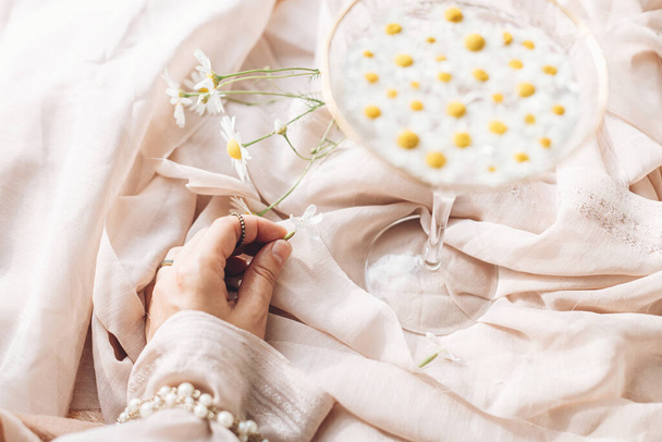Hand with jewelry holding white flower on background of soft beige fabric with stylish wineglass with daisy flowers in water. Tender floral aesthetic. Creative summer image. Bohemian mood - 写真・画像