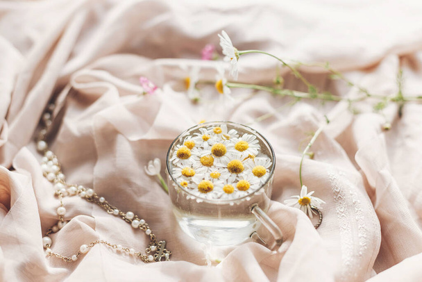 Tender floral aesthetic. Daisy flowers in water in glass cup on background of soft beige fabric with wildflowers and jewelry. Creative summer image with space for text. Bohemian mood - Zdjęcie, obraz