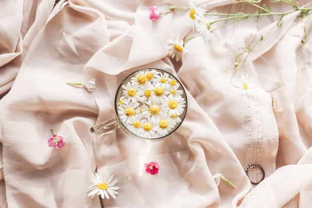 Tender floral aesthetic. Daisy flowers in water in glass cup on background of soft beige fabric with wildflowers and jewelry. Creative summer image, flat lay. Bohemian mood - Photo, image