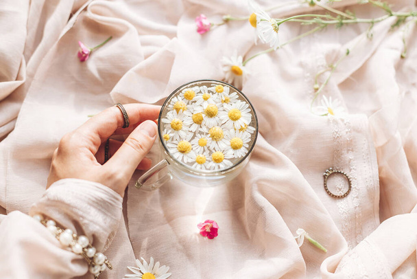 Mano con portagioie tazza di vetro con fiori di margherita in acqua su sfondo di morbido tessuto beige con fiori di campo. Tenera estetica floreale. Immagine creativa estiva, posa piatta. Umore boemo - Foto, immagini