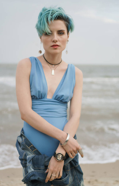 Fiatal lány kék fürdőruhában farmerdzsekiben a csípőjén pózol a tengerparton. - Fotó, kép