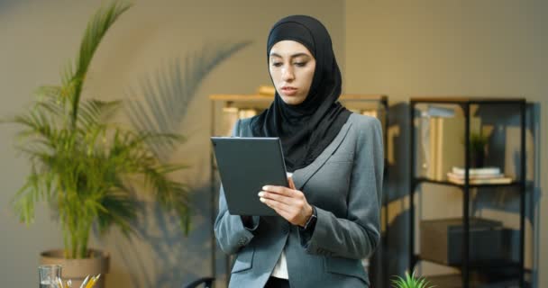 Portrait of Arabian beautiful woman in black hijab standing in office, tapping and scrolling on tablet device. Muslim happy businesswoman in headscarf smiling to camera. Arab female using computer - Séquence, vidéo
