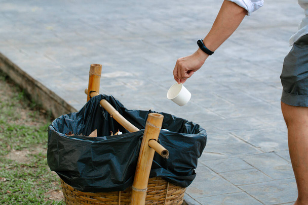 Facet wyrzucający papierowy kubek do kosza na śmieci na zewnątrz. Koncepcja recyklingu w słoneczny dzień na tle zielonej trawy. - Zdjęcie, obraz