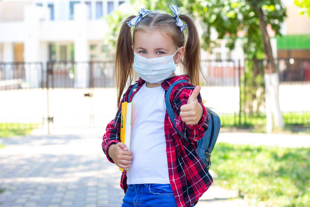 happy schoolgirl in a protective mask with a backpack and a textbook in her hands. in a white T-shirt and a plaid shirt - Фото, изображение