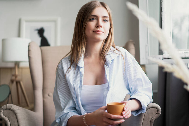 Enjoying her fresh coffee. Beautiful young woman drinking coffee and looking through window while sitting at armchair at home - Foto, afbeelding