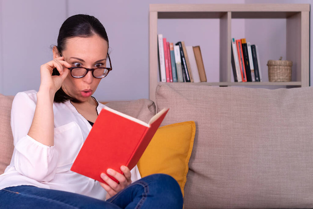 Pretty young brunette woman shocked reading a book sitting on the sofa at home. She is wearing a white shirt, glasses and a ponytail. - Photo, Image