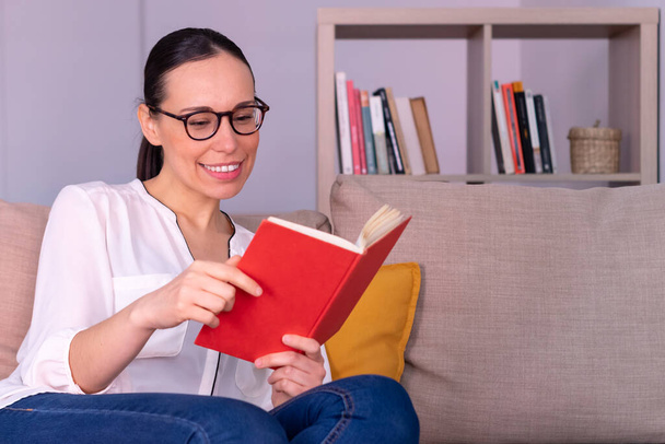 Ładna, młoda brunetka uśmiecha się czytając książkę siedzącą na kanapie w domu. Ma białą koszulę, okulary i kucyk.. - Zdjęcie, obraz