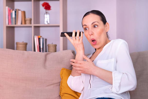 Surprised pretty young brunette woman listening to an audio on the smartphone sitting on the sofa at home. She is wearing a white shirt and a ponytail. - 写真・画像
