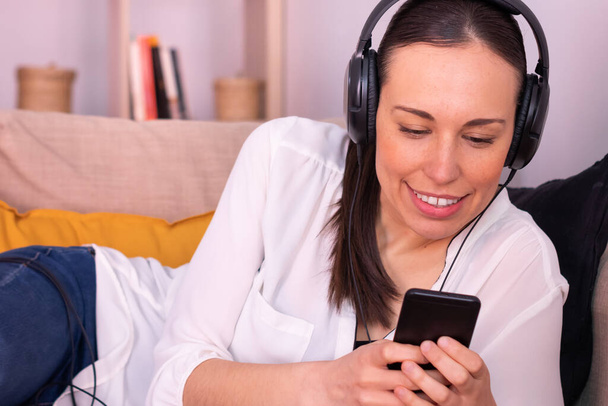 Kaunis brunette nuori nainen hymyilee ojensi sohvalla kotona älypuhelimella ja käyttää kuulokkeita. - Valokuva, kuva