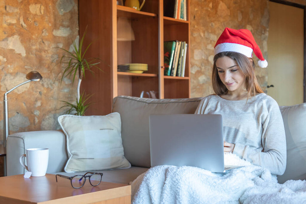 Noel Baba şapkalı gülümseyen genç bir kadın evinde dizüstü bilgisayarın önünde oturmuş film seyrediyor. - Fotoğraf, Görsel