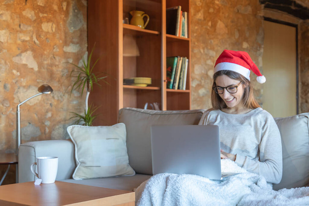 Šťastná mladá žena chatuje s brýlemi a Santa klobouk sedí na pohovce doma před notebookem - Fotografie, Obrázek
