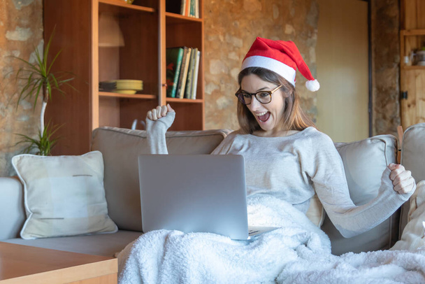 Евфорична жінка посміхається руками витягнутими окулярами і Санта-капелюх сидить на дивані вдома перед ноутбуком
 - Фото, зображення