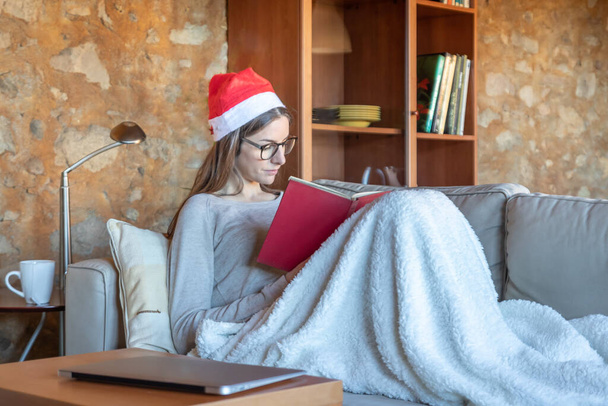 Konzentrierte hübsche brünette junge Frau, die zu Hause auf dem Sofa sitzt und mit Weihnachtsmütze ein Buch liest, das mit einer Decke bedeckt ist. Sie trägt eine Brille. - Foto, Bild