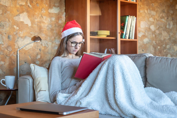 Triste jolie jeune femme brune assise sur le canapé à la maison avec le chapeau du Père Noël lisant un livre couvert d'une couverture. Elle porte des lunettes.. - Photo, image