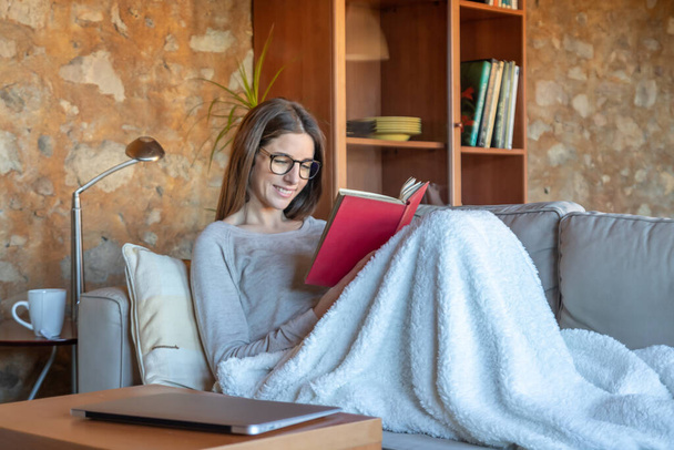 Uma jovem bonita sorrindo sentada no sofá em casa lendo um livro. Ela está usando óculos e cabelos castanhos longos. Imagem horizontal
 - Foto, Imagem