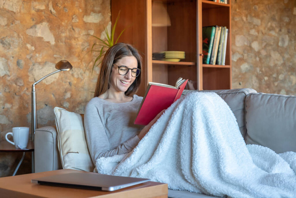 Pretty young woman laughing sitting on the sofa at home reading a book. She is wearing glasses and long brown hair. Horizontal image - Foto, afbeelding