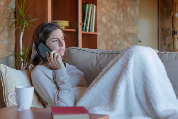Jeune femme brune assez sérieuse parlant sur le smartphone assis sur le canapé à la maison couvert d'une couverture regardant vers l'horizon. - Photo, image