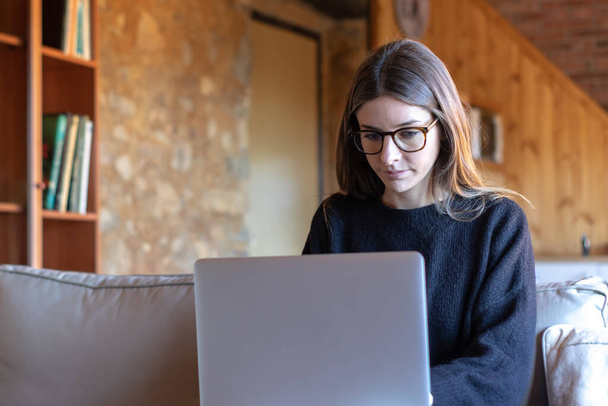 Giovane donna seria con occhiali da vista che scrive davanti al computer portatile seduto sul divano di casa - Foto, immagini