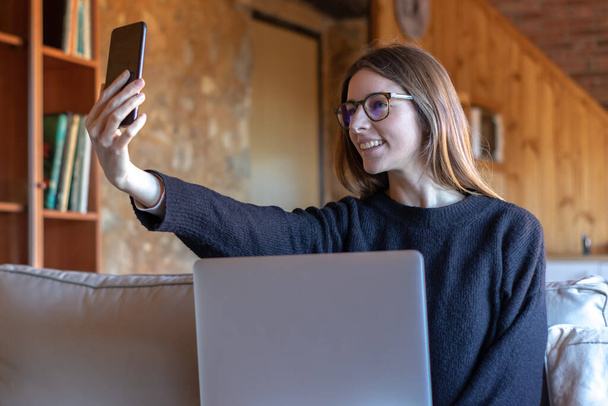 Молодая брюнетка делает видеозвонок со смартфоном перед ноутбуком, сидя дома на диване в черном свитере и очках - Фото, изображение