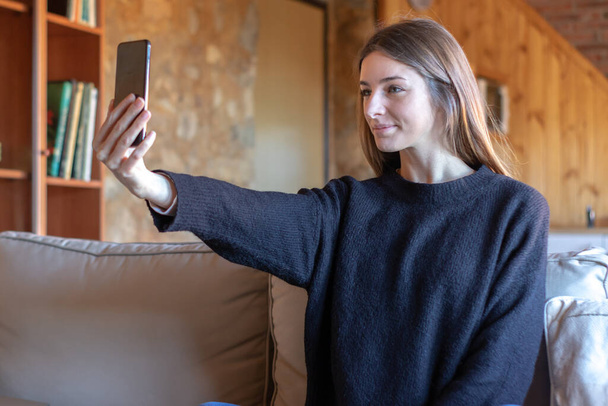 Mladý pěkný brunetka žena přičemž selfie s smartphone sedí na pohovce doma nosí černý svetr - Fotografie, Obrázek